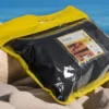 Emballage transparent avec bordure jaune de l'oreiller Litex, étiquette "Voyage" affichée, placée sur des rochers avec l'océan en arrière-plan.