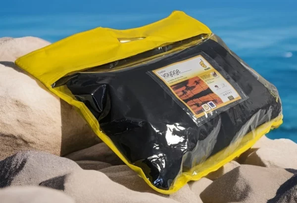 Emballage transparent avec bordure jaune de l'oreiller Litex, étiquette "Voyage" affichée, placée sur des rochers avec l'océan en arrière-plan.