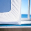 Protège-matelas blanc ajustable Litex avec élastique bleu, vue partielle avec arrière-plan marin.
