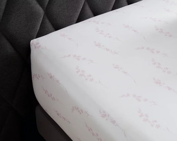 Un coin de drap plat en coton bio orné de motifs floraux roses posé sur un lit avec tête de lit en tissu gris foncé.