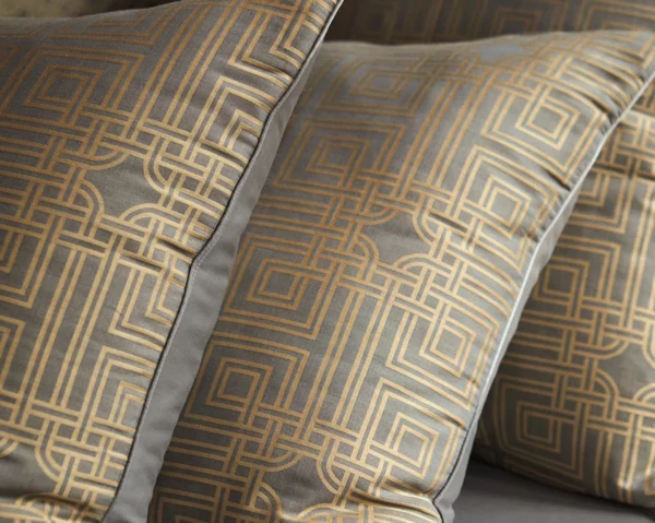 Deux taies d'oreillers de la collection Trianon affichant des motifs géométriques dorés sur fond gris, bordées d’un liseré élégant.