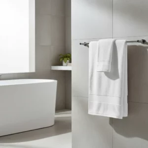 Drap de bain et serviette Pétale Jasmin blancs suspendus à un porte-serviette dans une salle de bain moderne avec carrelage gris et baignoire.