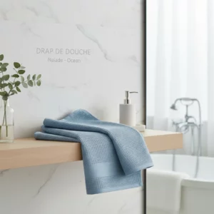 Drap de douche Naïade Ocean Anne de Solène plié sur une étagère en bois, décorant une salle de bain moderne aux murs de marbre blanc.