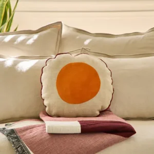 Coussin rond Bérénice Anne de Solène posé sur des oreillers de lit en lin clair avec un plaid bordeaux.