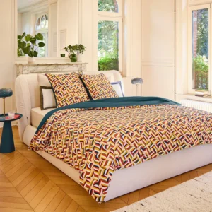 Lit de luxe habillé de la parure de couette Claudia en satin de coton bio, vue d'angle dans une chambre lumineuse avec parquet et cheminée.